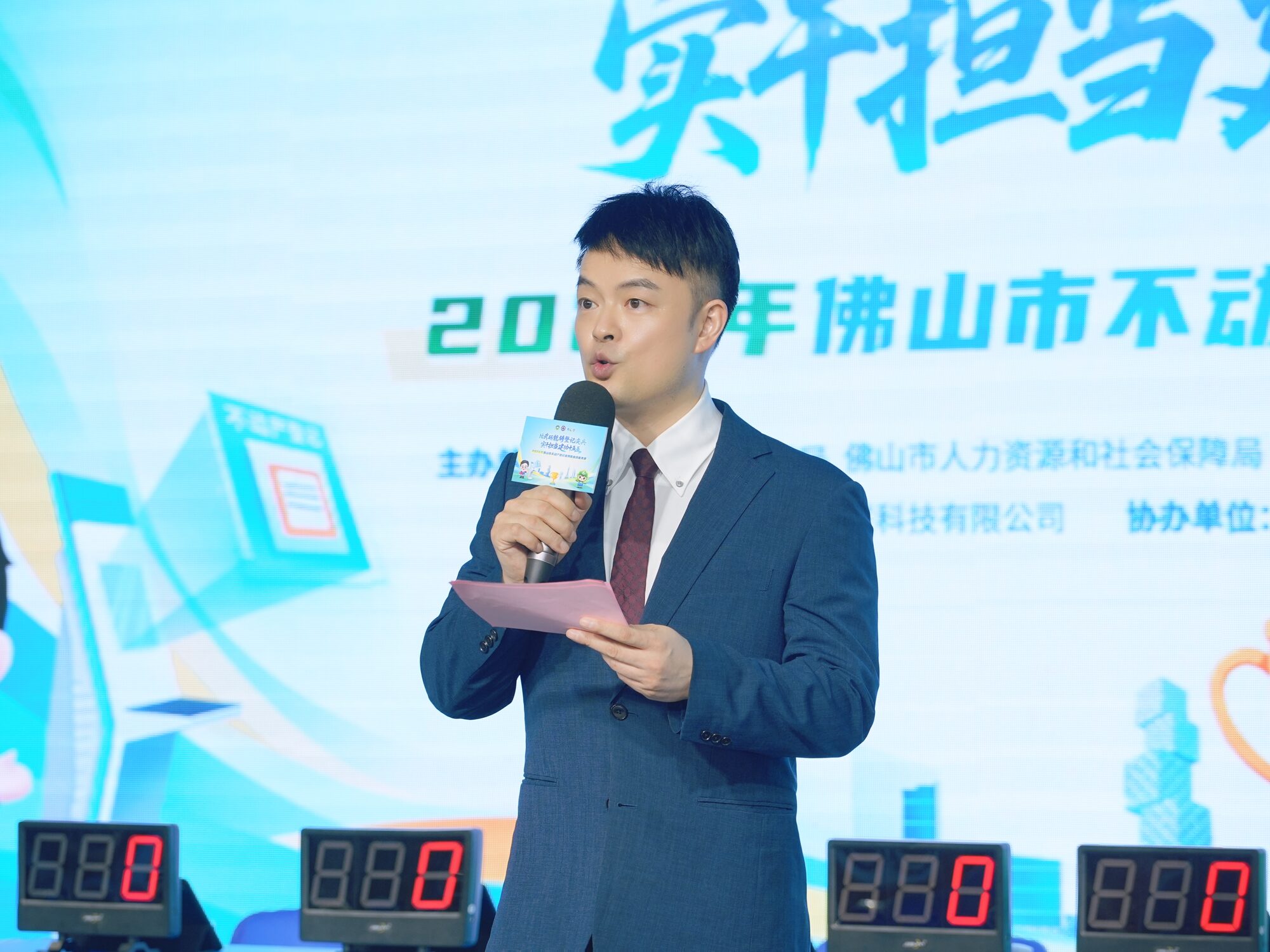 常安在四台计分屏前主持计分与风采展示环节，台上大屏显示主办、协办单位标识